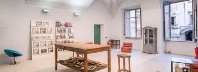 Sala interna ai Magazzini fotografici con grande tavolo in legno, sedie di vari stili e due ampie finestre.
